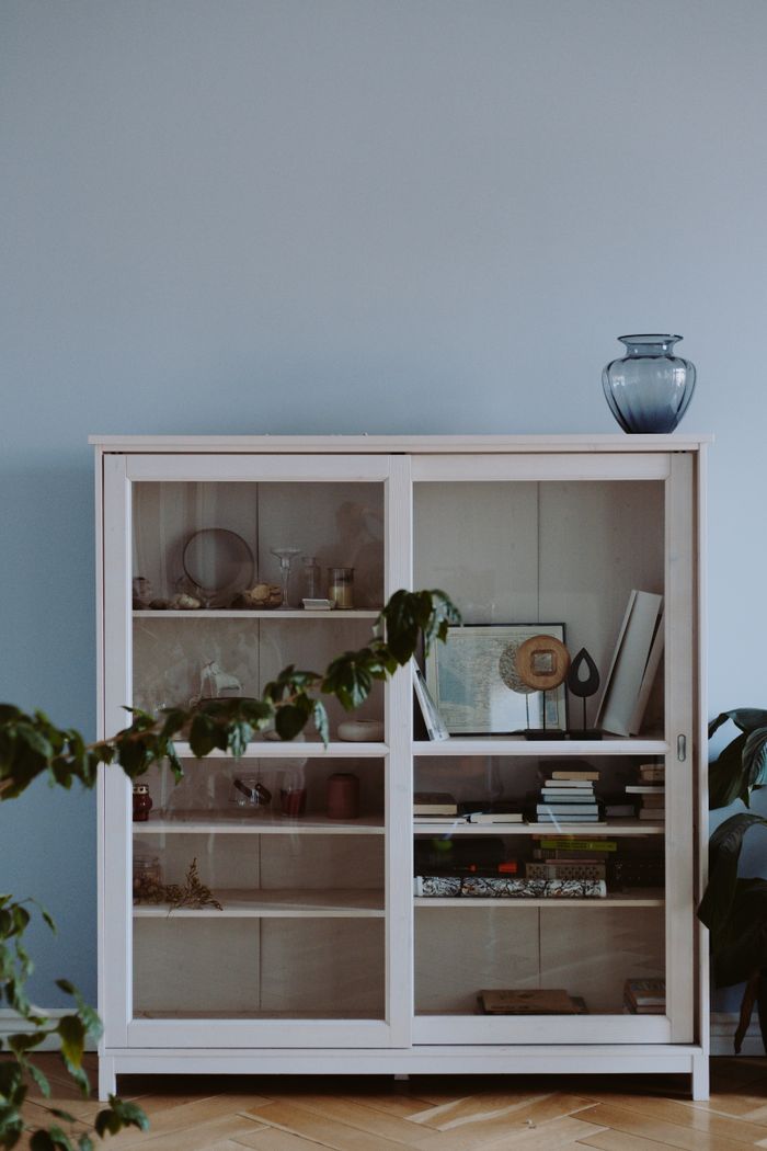 A wooden glass cabinet with shelves filled with books and decorative items, set against a light blue wall.